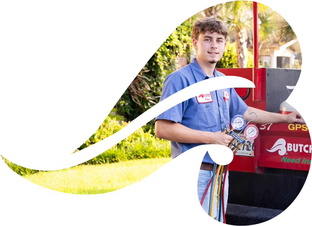Young HVAC technician in blue uniform holding manifold gauges by a red service truck at a residential property