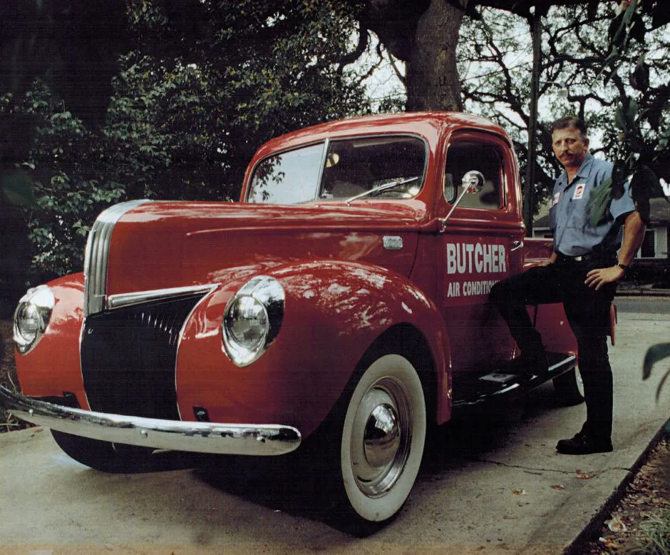 Man in work shirt leaning on classic red pickup truck labeled Butcher Air Conditioning, parked under trees