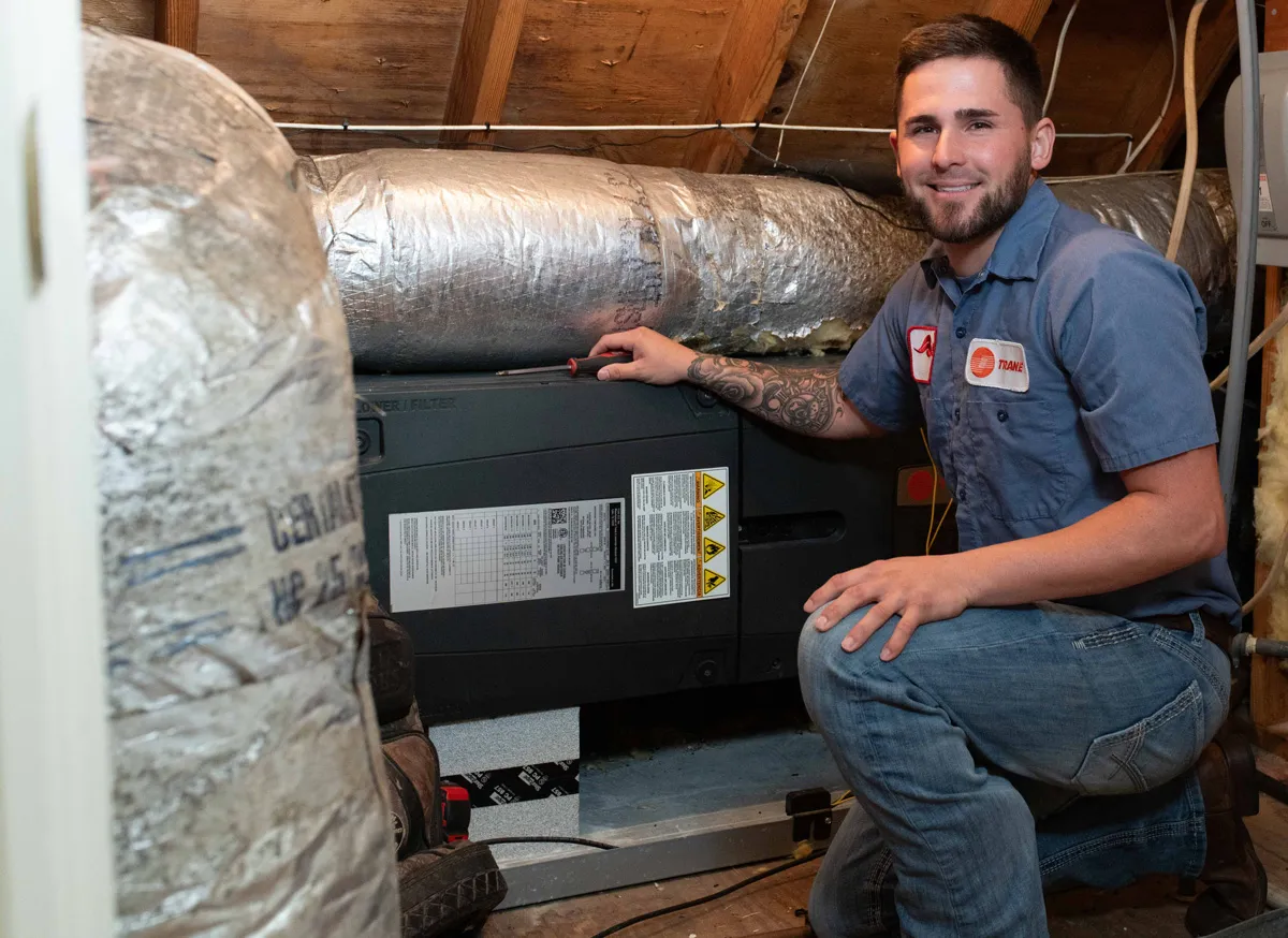 A man HVAC technician kneeling in attic inspecting furnace unit, smiling in blue uniform, ductwork and insulation visible