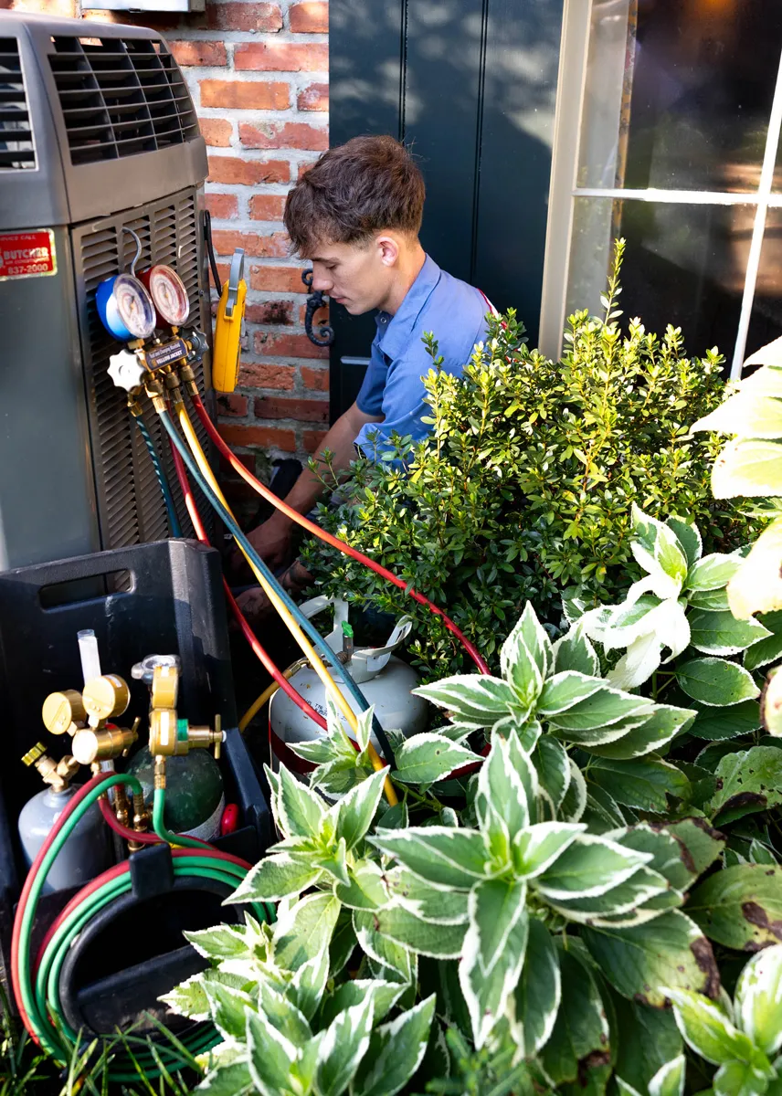 A man servicing an outdoor HVAC unit with manifold gauges, hoses and refrigerant tanks beside home landscaping