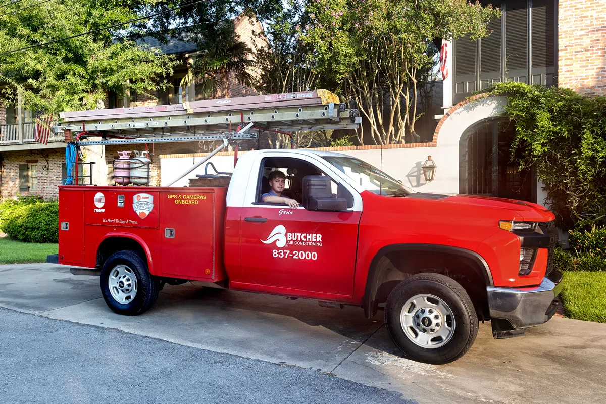 Red Butcher Air Conditioning service truck with technician at wheel and ladders on roof parked in residential driveway