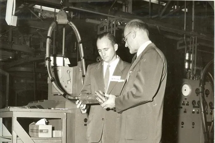 Two men in suits inspecting a metal part on a factory floor amid industrial machinery and control panels.