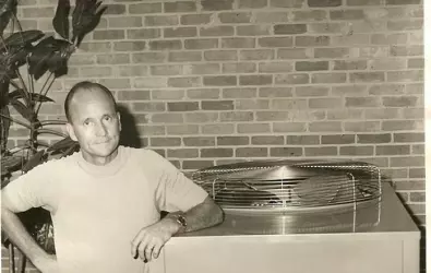 Black-and-white photo of a man leaning on a vintage industrial air conditioning unit by a brick wall