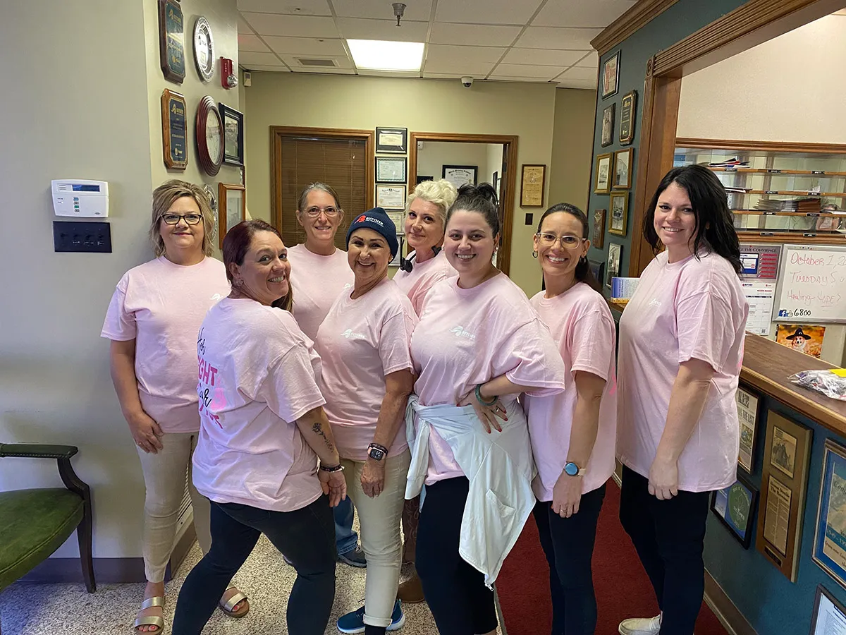 Group of eight women in pink shirts smiling inside an office, team photo for breast cancer awareness.