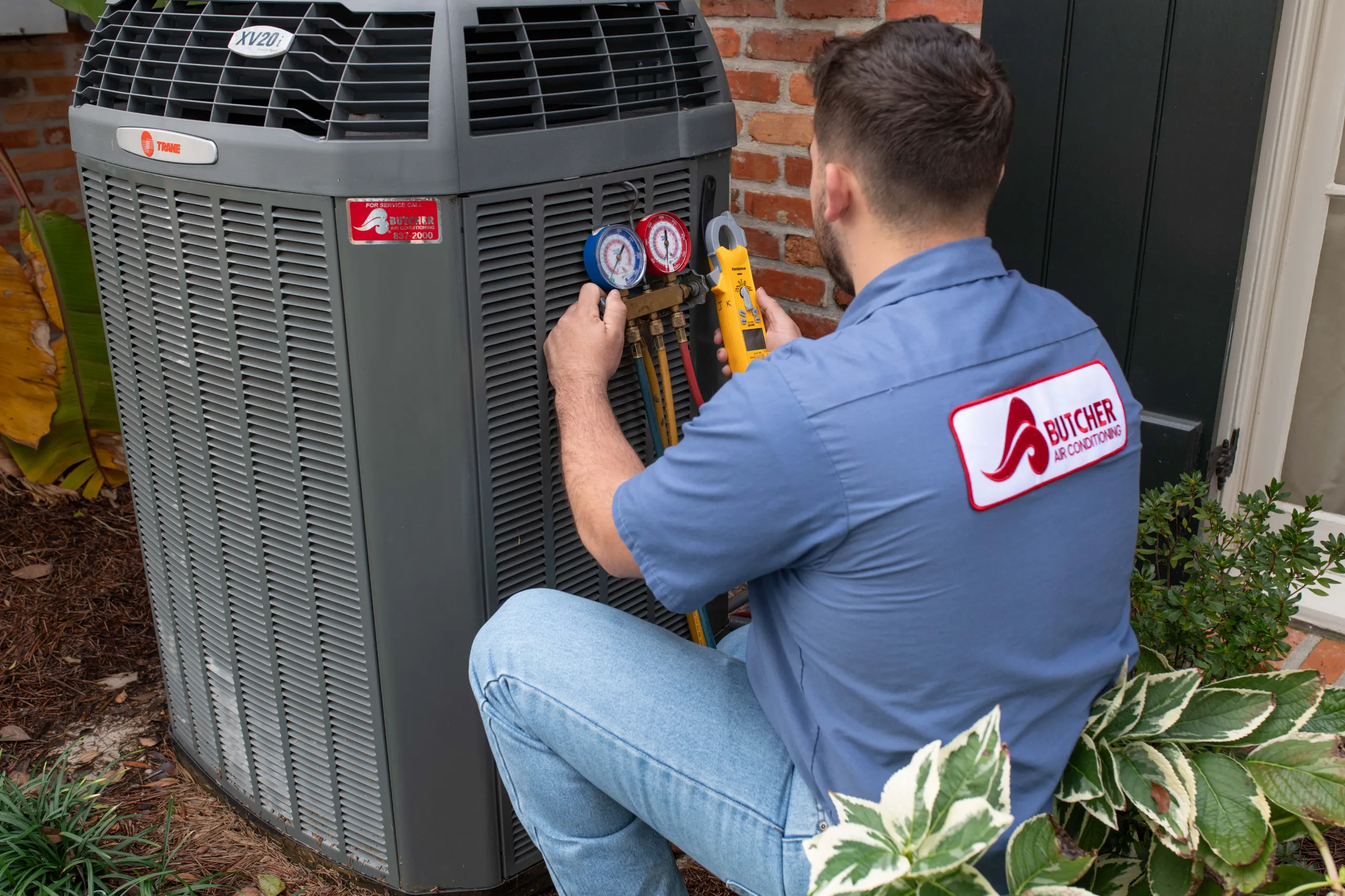 A man performing HVAC maintenance on a residential air conditioner using manifold gauges and clamp meter