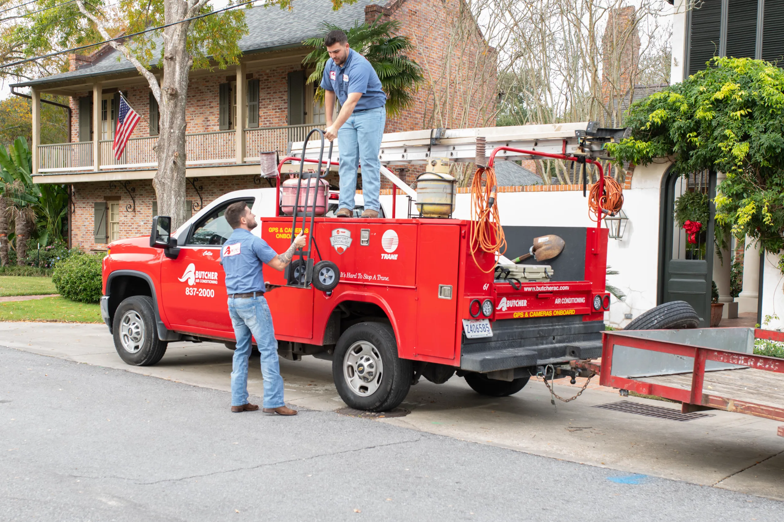 Two HVAC technicians loading a ladder onto a red service truck in front of a brick residential house
