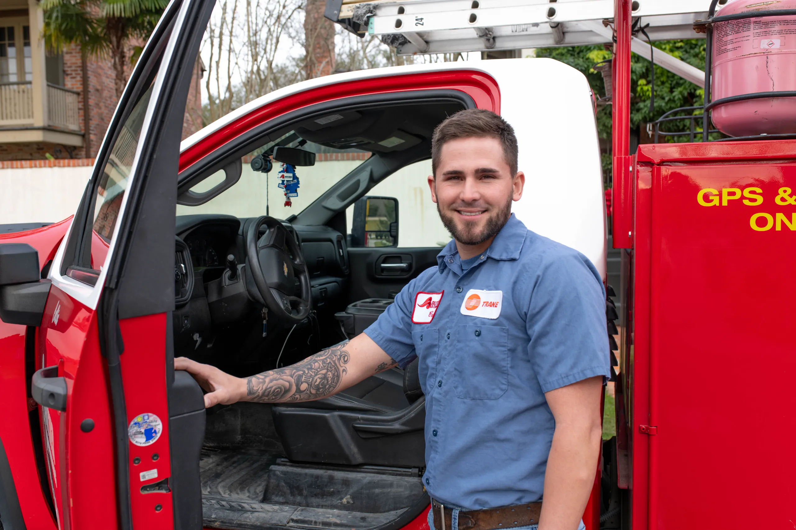 Smiling man in technician uniform with tattooed forearm standing by red service truck with ladder equipment