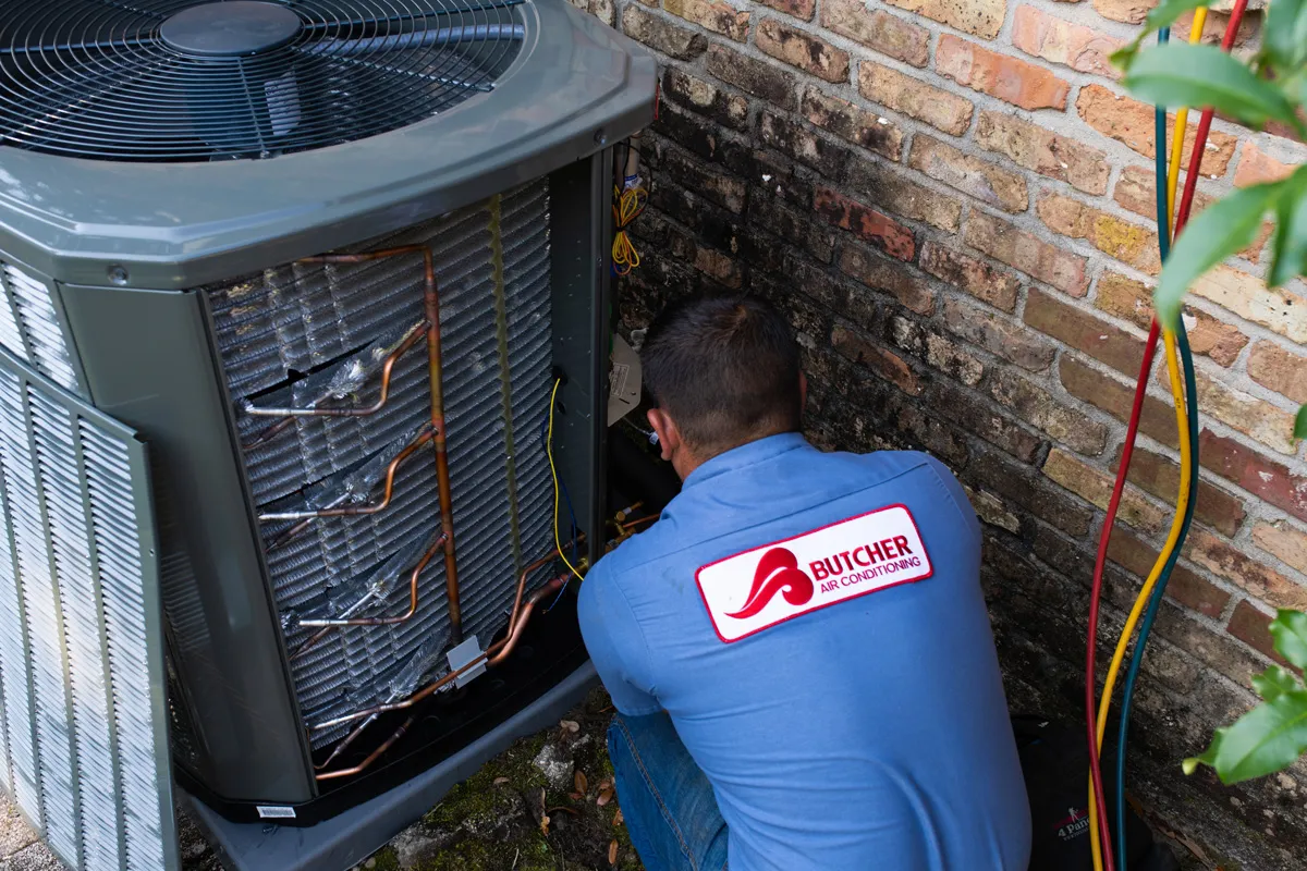 HVAC technician performing AC repair on outdoor air conditioning unit with refrigerant hoses and tools by brick wall