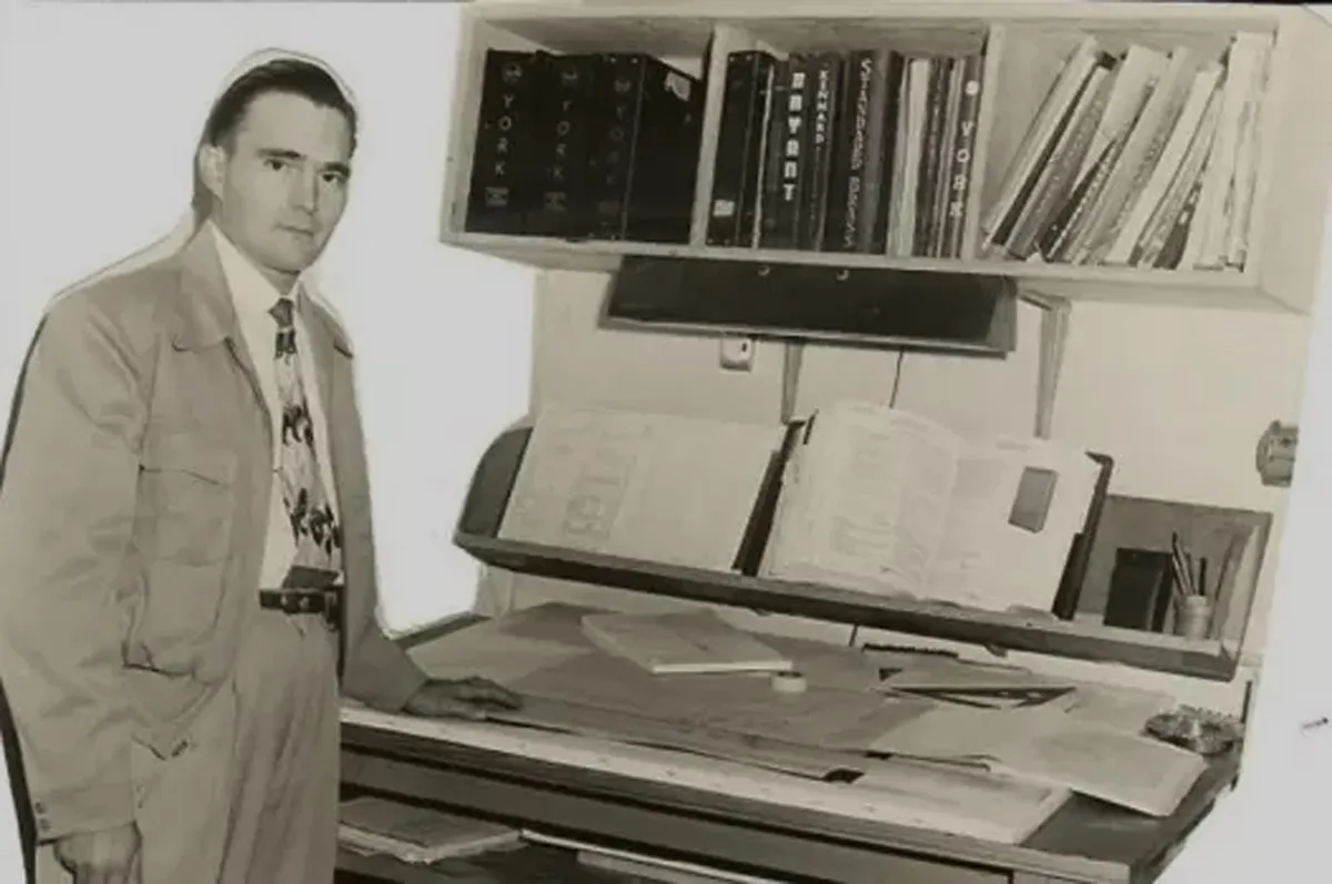 Black-and-white photo of a man at a drafting table with architectural plans and open reference books on shelf