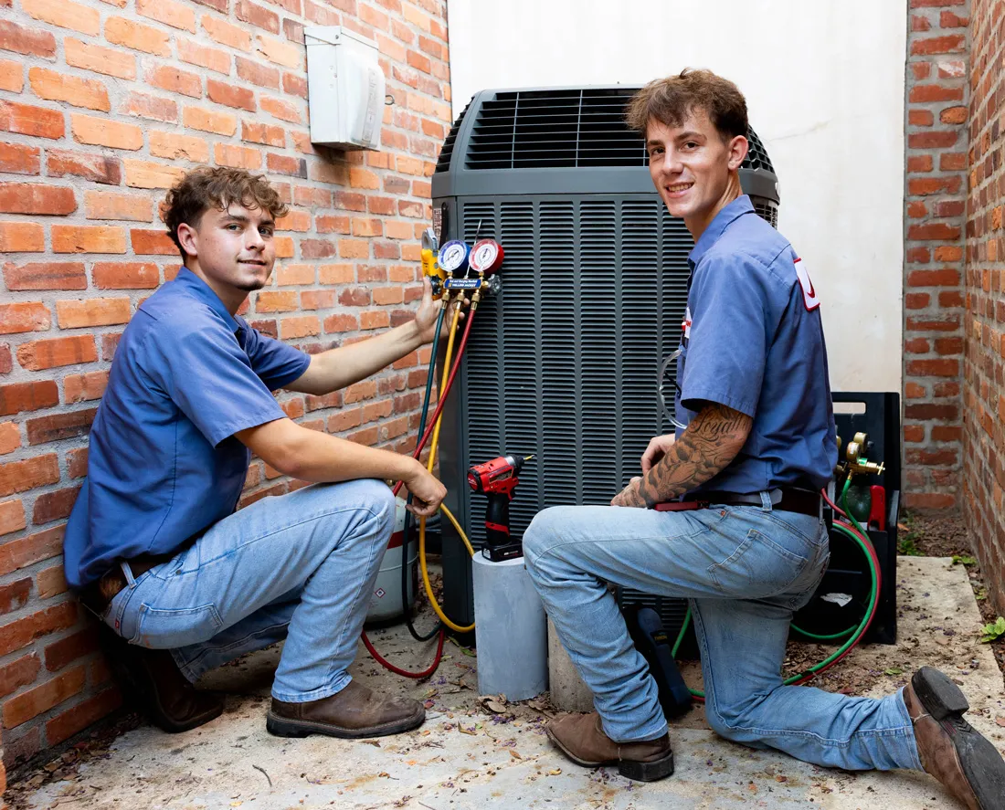 Two HVAC technicians kneeling beside an outdoor air conditioning unit, connecting gauges and tools for maintenance