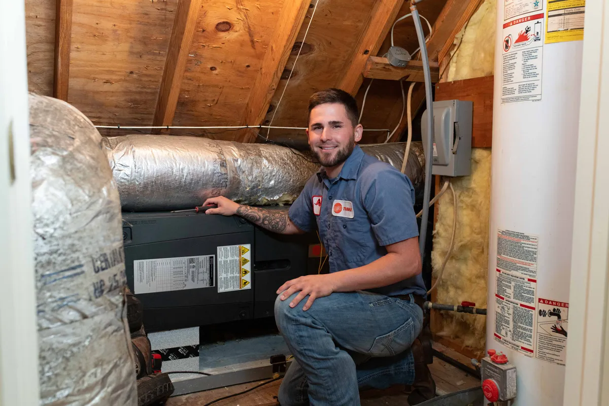 HVAC technician kneeling in attic inspecting air handler near water heater, wearing blue uniform.