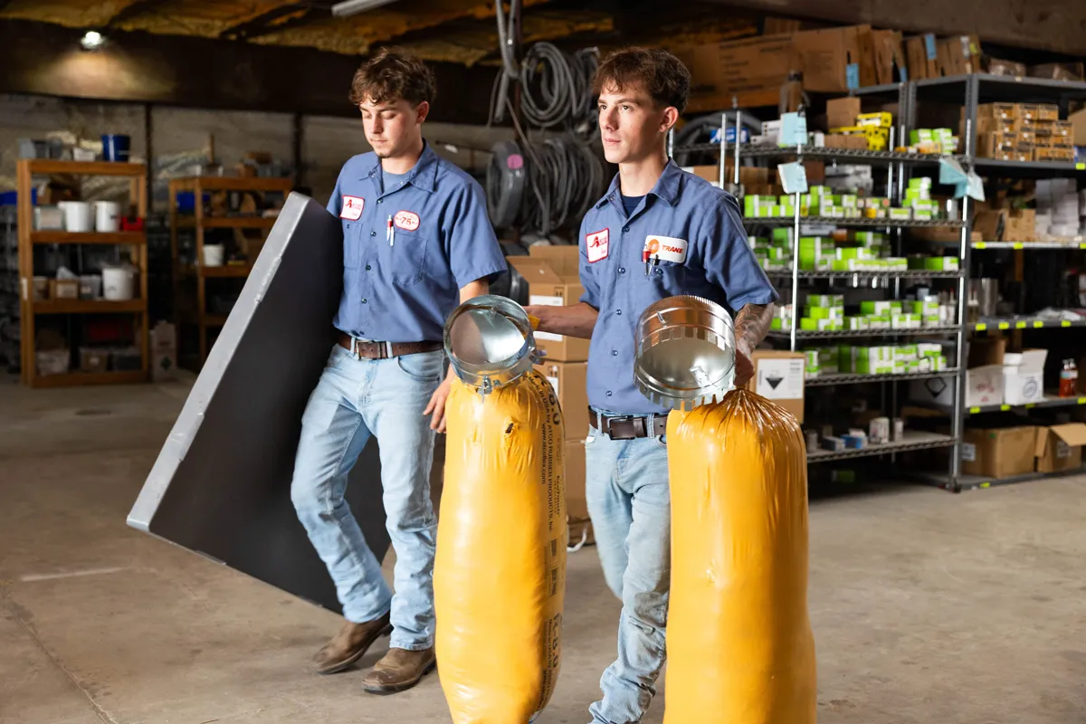 Two HVAC technicians carrying duct collars and yellow insulation rolls in a warehouse supply room