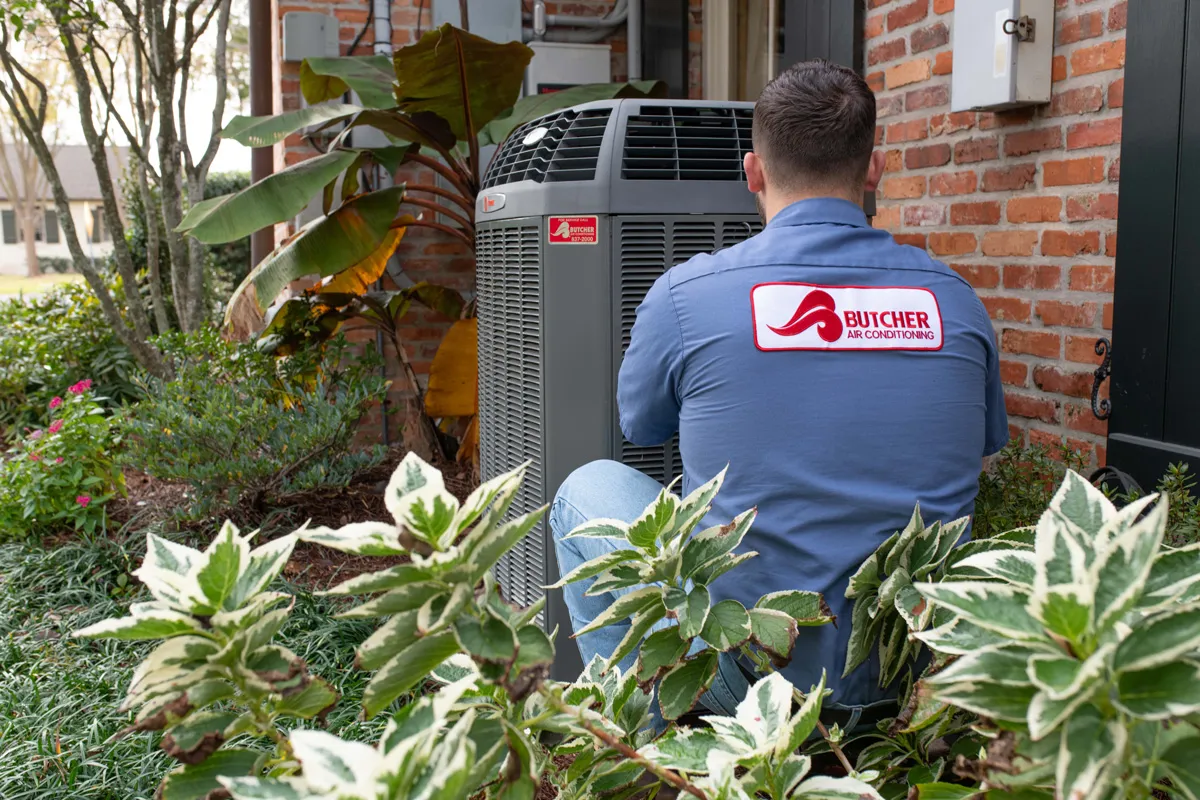 A man in Butcher Air Conditioning uniform servicing an outdoor air conditioner beside a brick house