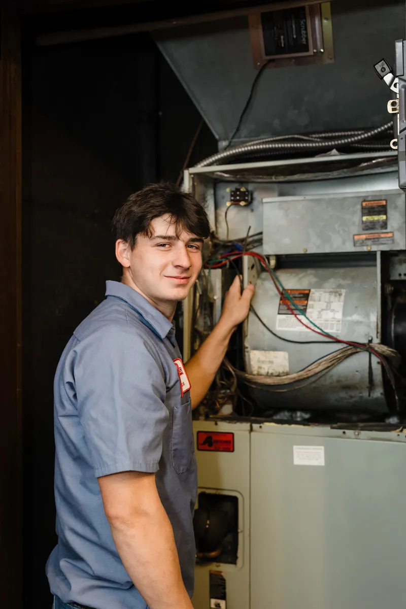 HVAC technician inspecting furnace wiring in utility room, a smiling man in blue work shirt