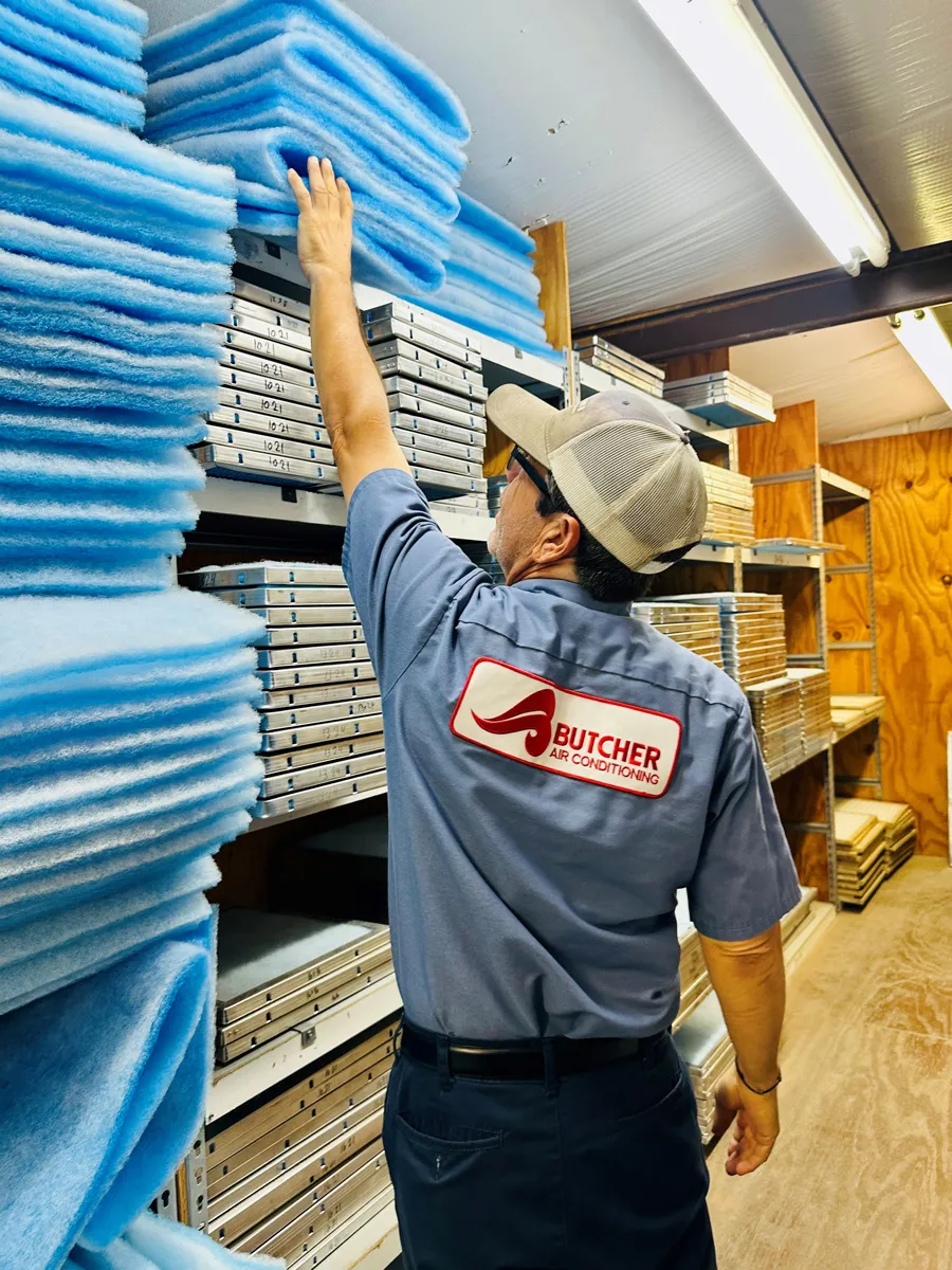 A man HVAC technician in Butcher Air Conditioning uniform reaching for blue air filter media on warehouse shelves