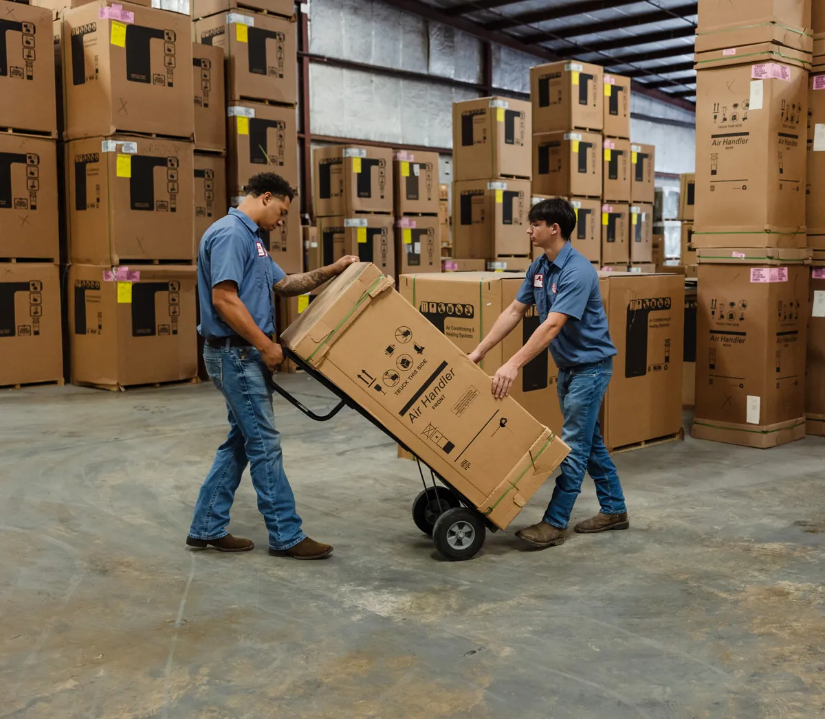 Two warehouse workers move a boxed air handler on a hand truck amid stacked boxes in a distribution center