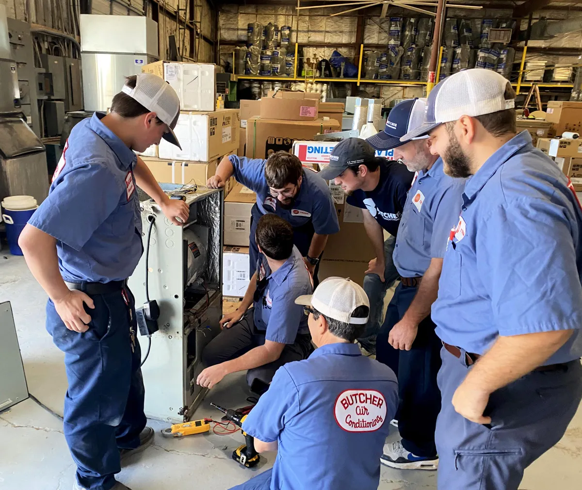 Group of HVAC technicians in blue uniforms repairing an air conditioning unit in a warehouse during hands-on training