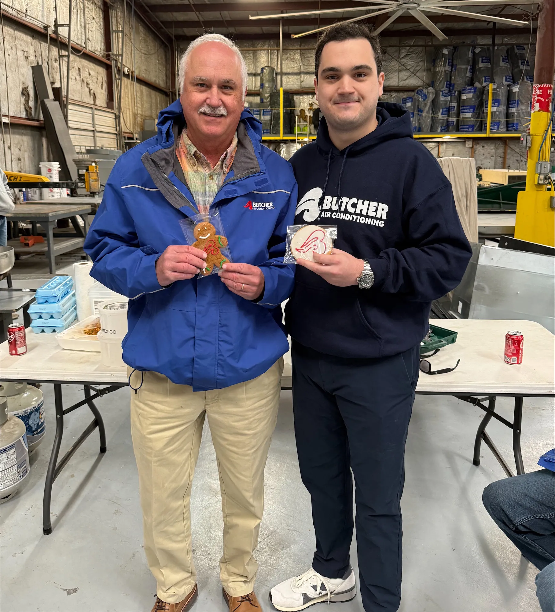 Two smiling men in Butcher Air Conditioning jackets holding cookies in a warehouse workshop