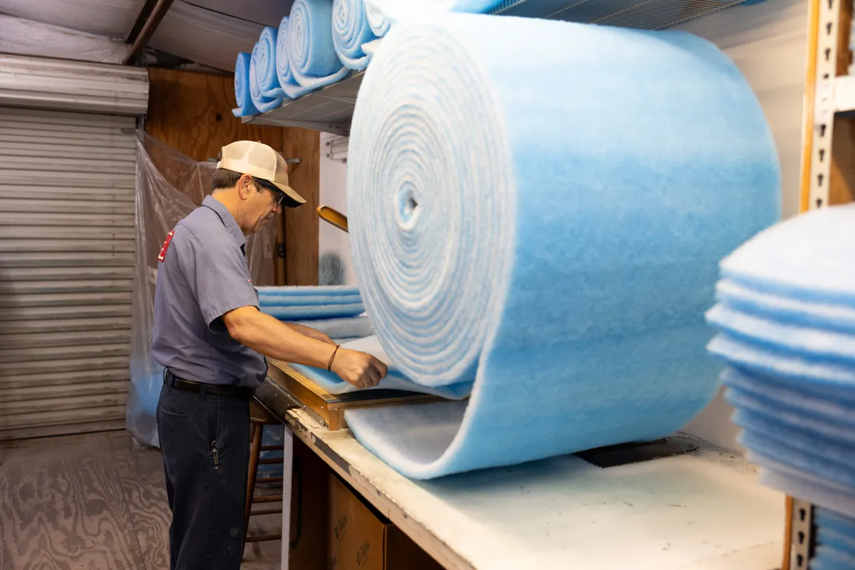 A man in a workshop unrolling and cutting large blue polyester batting roll for upholstery and insulation