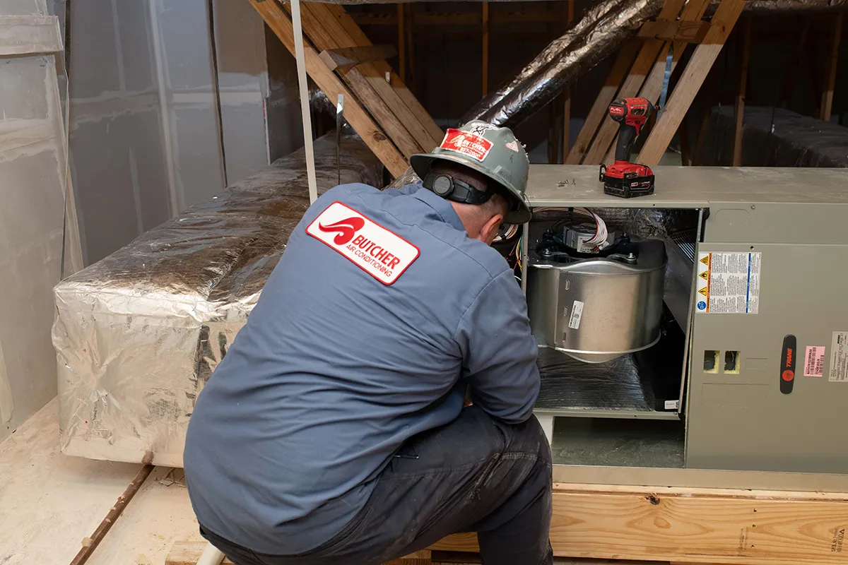 A man servicing HVAC air handler in attic, wearing Butcher Air Conditioning uniform and hard hat.