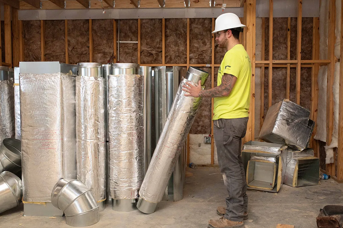 Construction worker installing insulated HVAC ductwork inside framed residential interior.