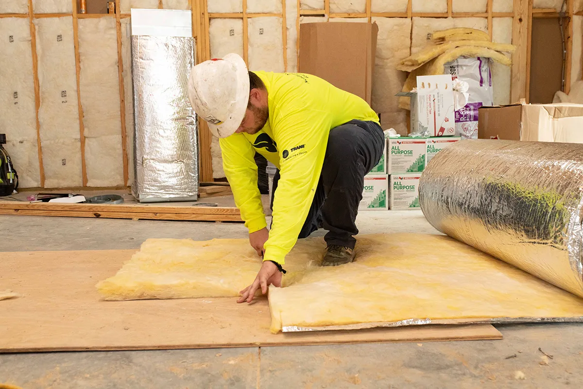 Construction worker in hard hat installing fiberglass insulation batt on subfloor during residential renovation