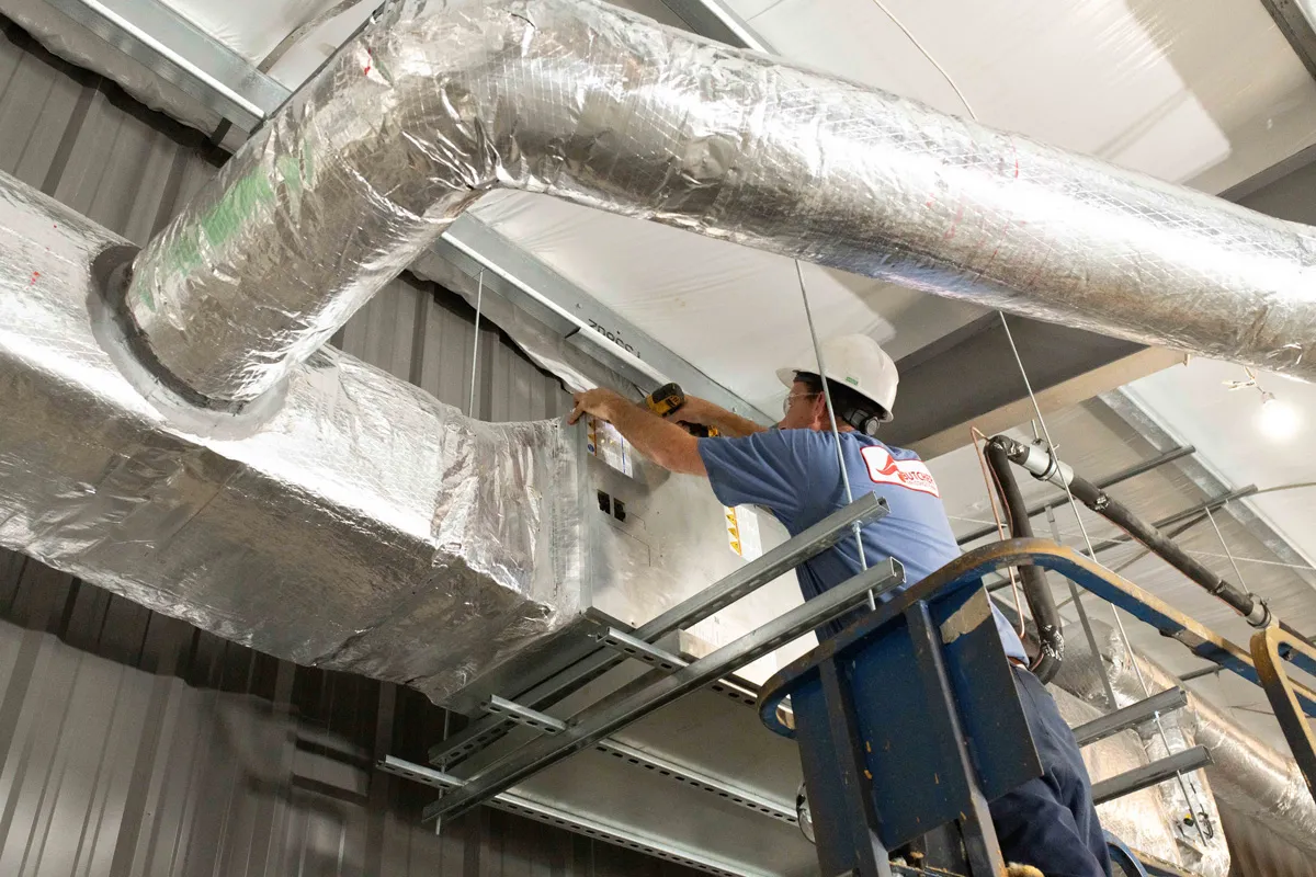 Commercial HVAC technician installing insulated ductwork on scissor lift in warehouse ceiling