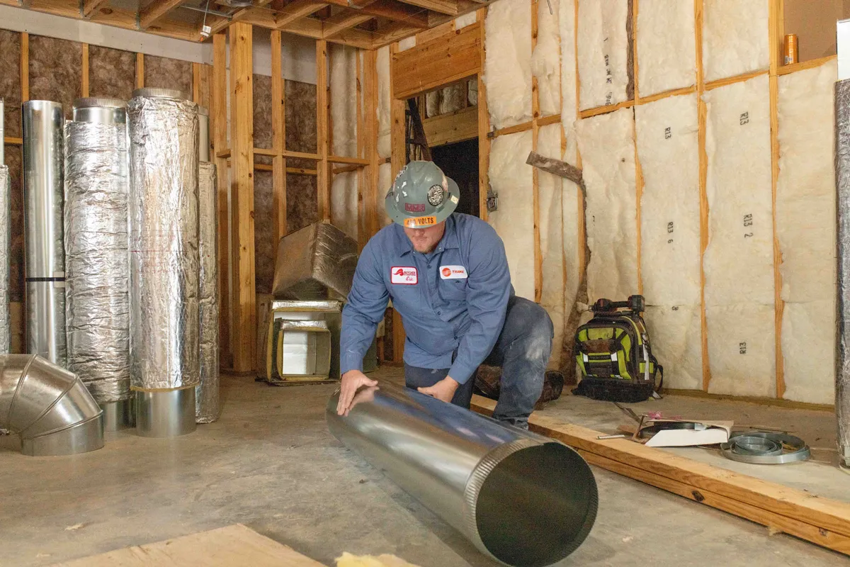 A man HVAC technician kneeling in framed room installing large metal ductwork beside wall insulation
