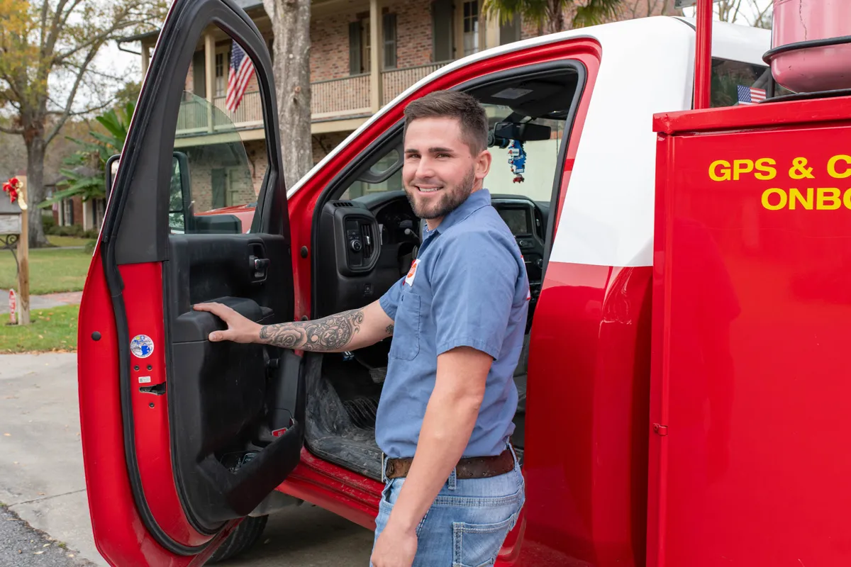 Smiling service technician in blue uniform with tattoo standing by open door of red service truck in residential neighborhood