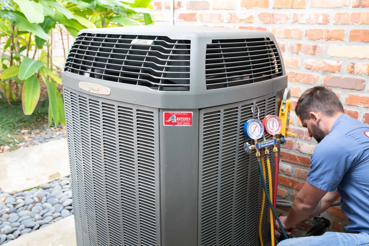 HVAC technician servicing outdoor air conditioning unit with manifold gauges and refrigerant hoses by brick wall