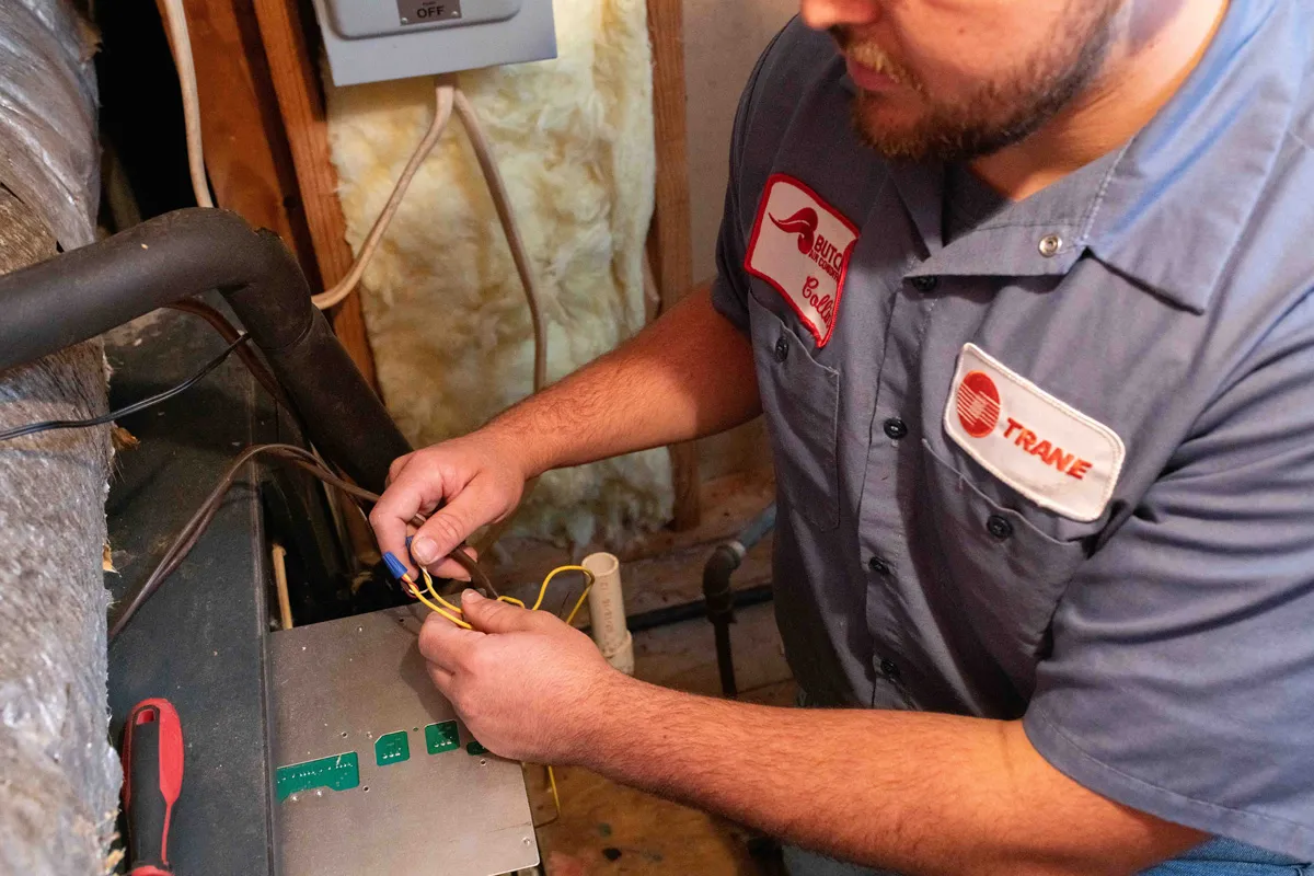 A man wiring a Trane furnace in a utility room, hands connecting yellow HVAC wires with a wire nut