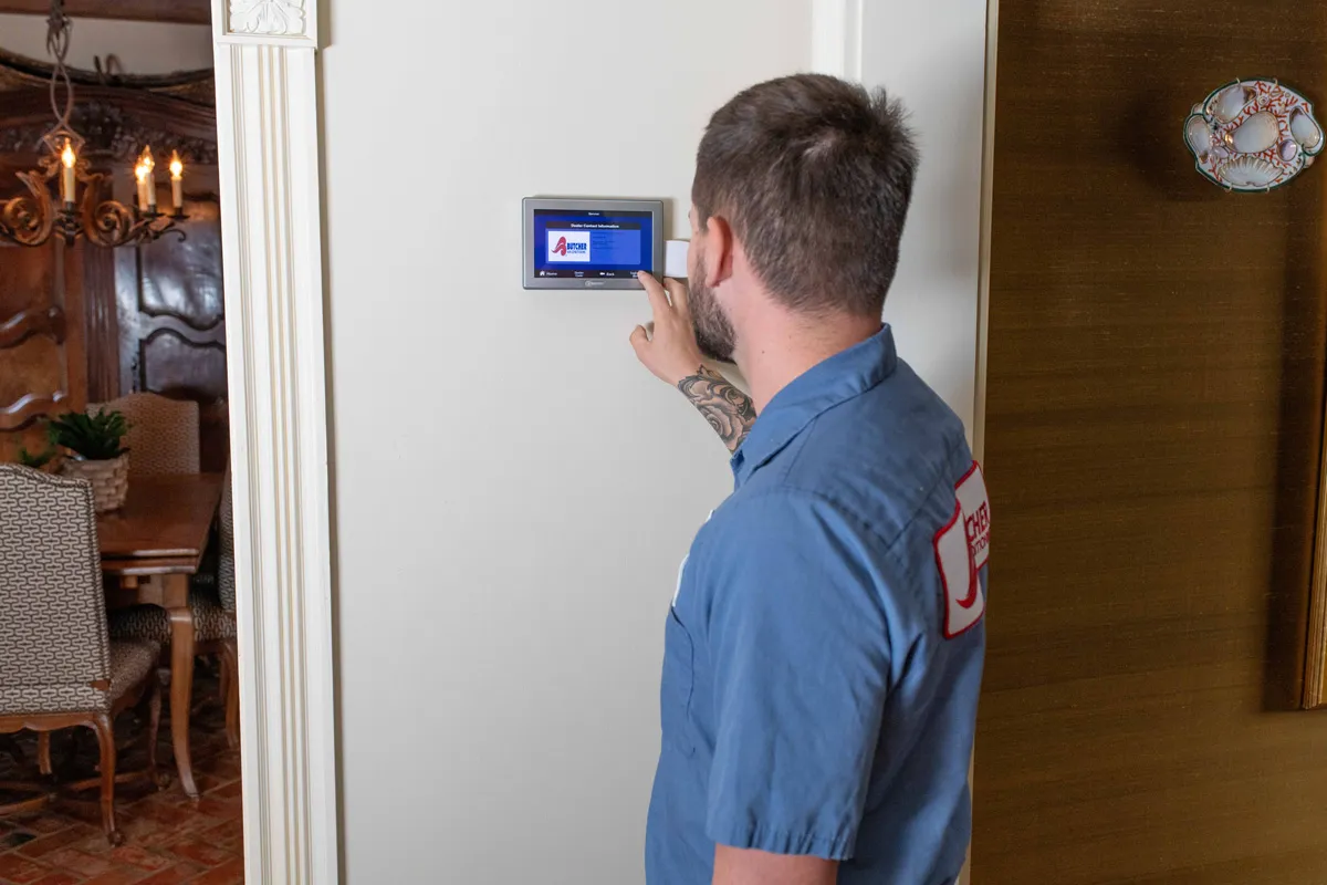 An HVAC technician, a man in a blue uniform, adjusts a smart thermostat touchscreen on a home wall