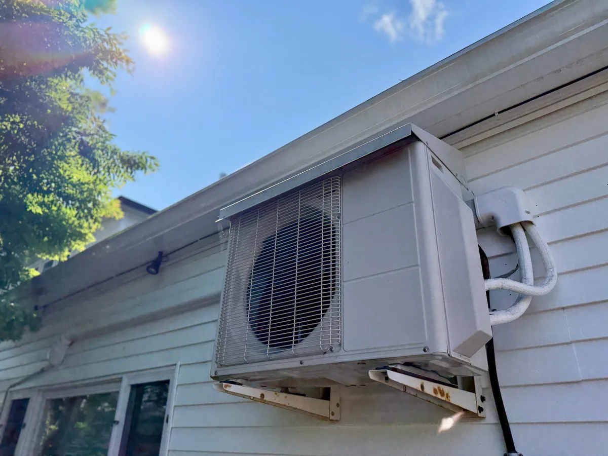 Wall-mounted outdoor mini-split AC unit on white house siding with insulated refrigerant lines under blue sky