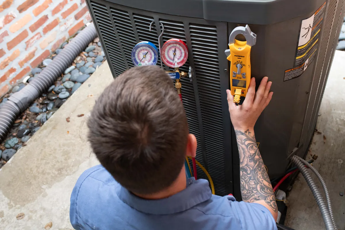 HVAC technician servicing outdoor air conditioner with manifold gauges and clamp meter, tattooed forearm visible