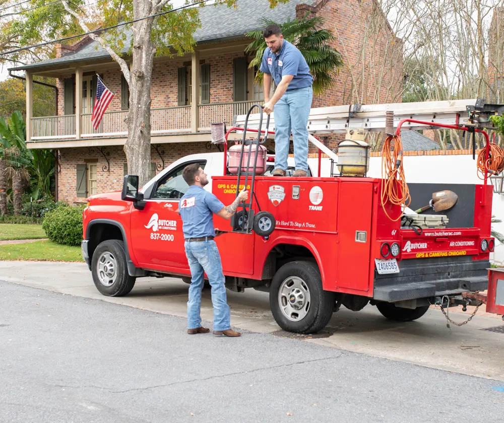 Two HVAC technicians loading equipment onto red service truck outside brick home with ladder and American flag