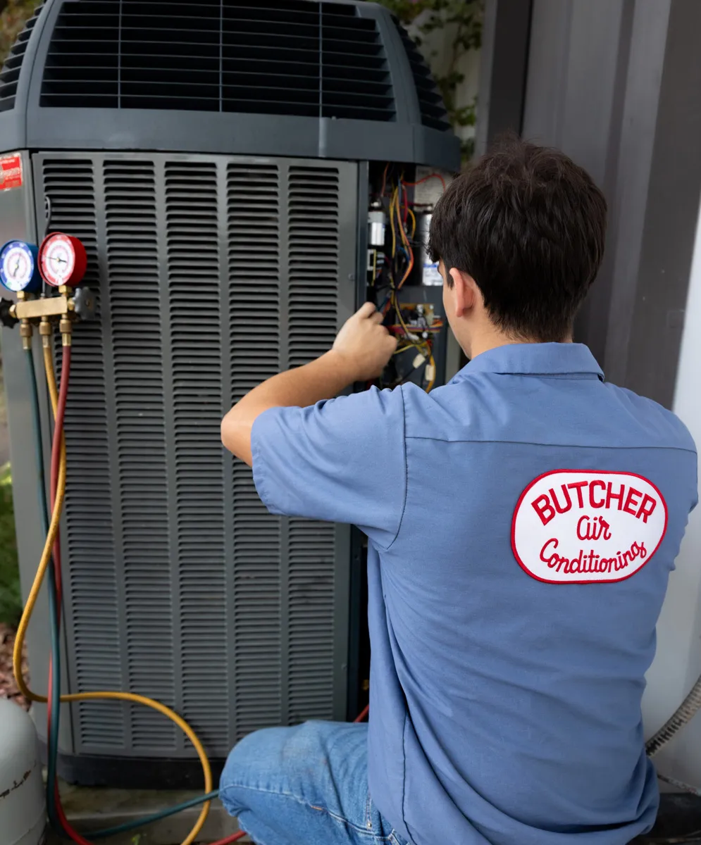 HVAC technician in blue uniform servicing an outdoor air conditioner condenser using manifold gauges and hoses