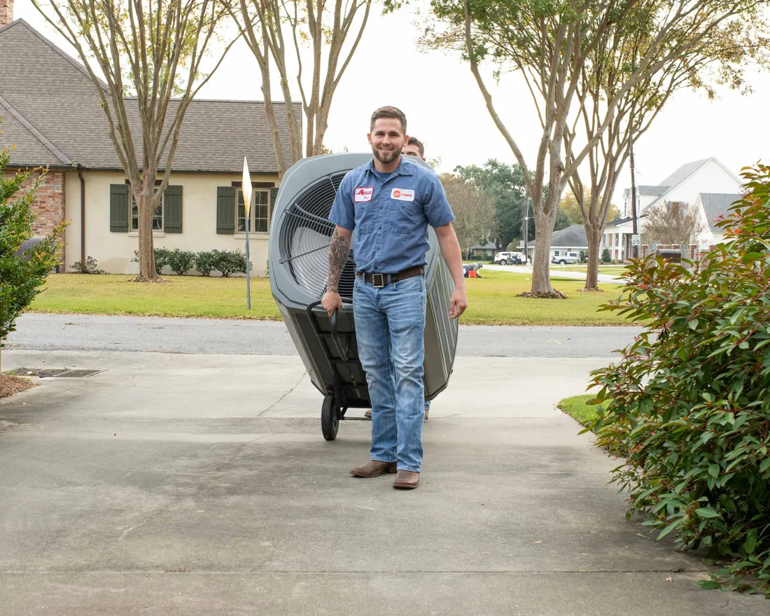HVAC technician carrying outdoor air conditioning unit on a dolly down a residential driveway, smiling man in uniform