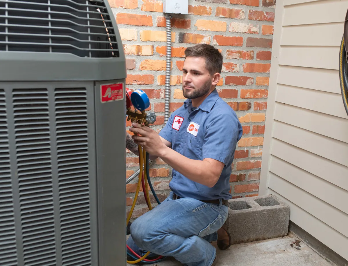 A man HVAC technician kneeling, checking gauges on outdoor AC unit beside brick wall for HVAC service
