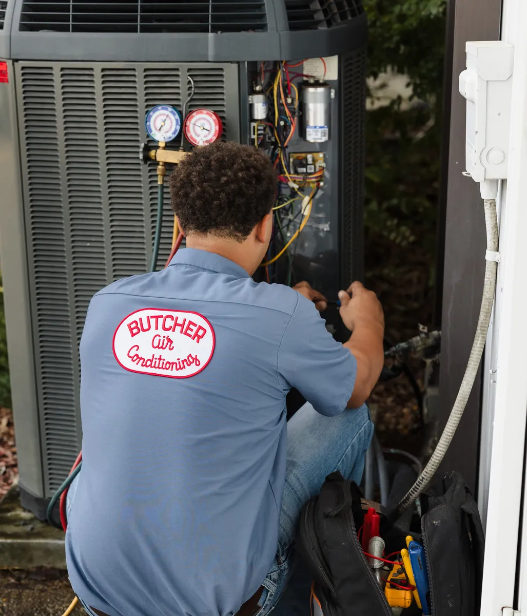 HVAC technician wearing Butcher Air Conditioning shirt repairing outdoor AC unit with pressure gauges and tools