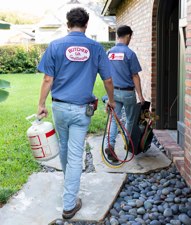 Two HVAC technicians in blue uniforms carrying a refrigerant tank and manifold with hoses into a house