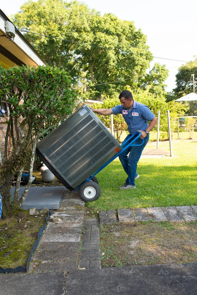 A man HVAC technician using hand truck to move an outdoor air conditioning unit in a backyard