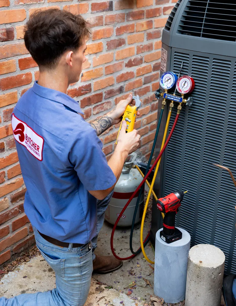 HVAC technician kneeling next to outdoor AC unit using manifold gauges and leak detector to service refrigerant lines