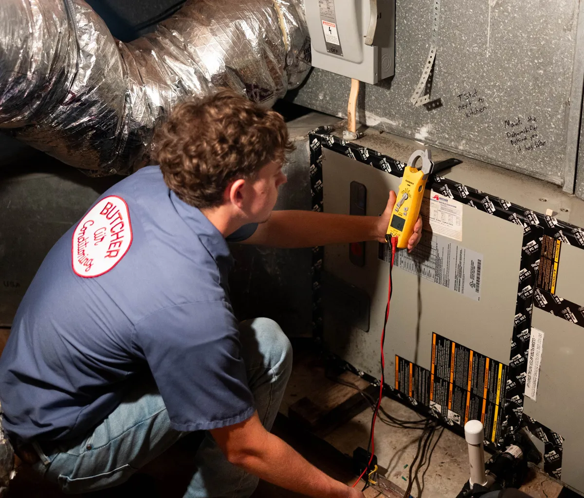 HVAC technician testing furnace wiring with yellow clamp meter in basement near ductwork