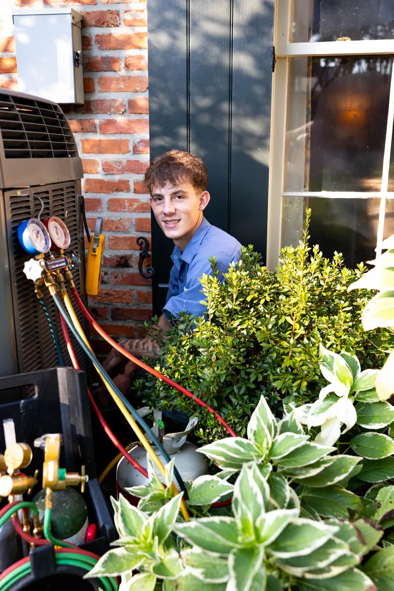 HVAC technician servicing outdoor air conditioner unit with gauges and hoses beside brick house and garden plants
