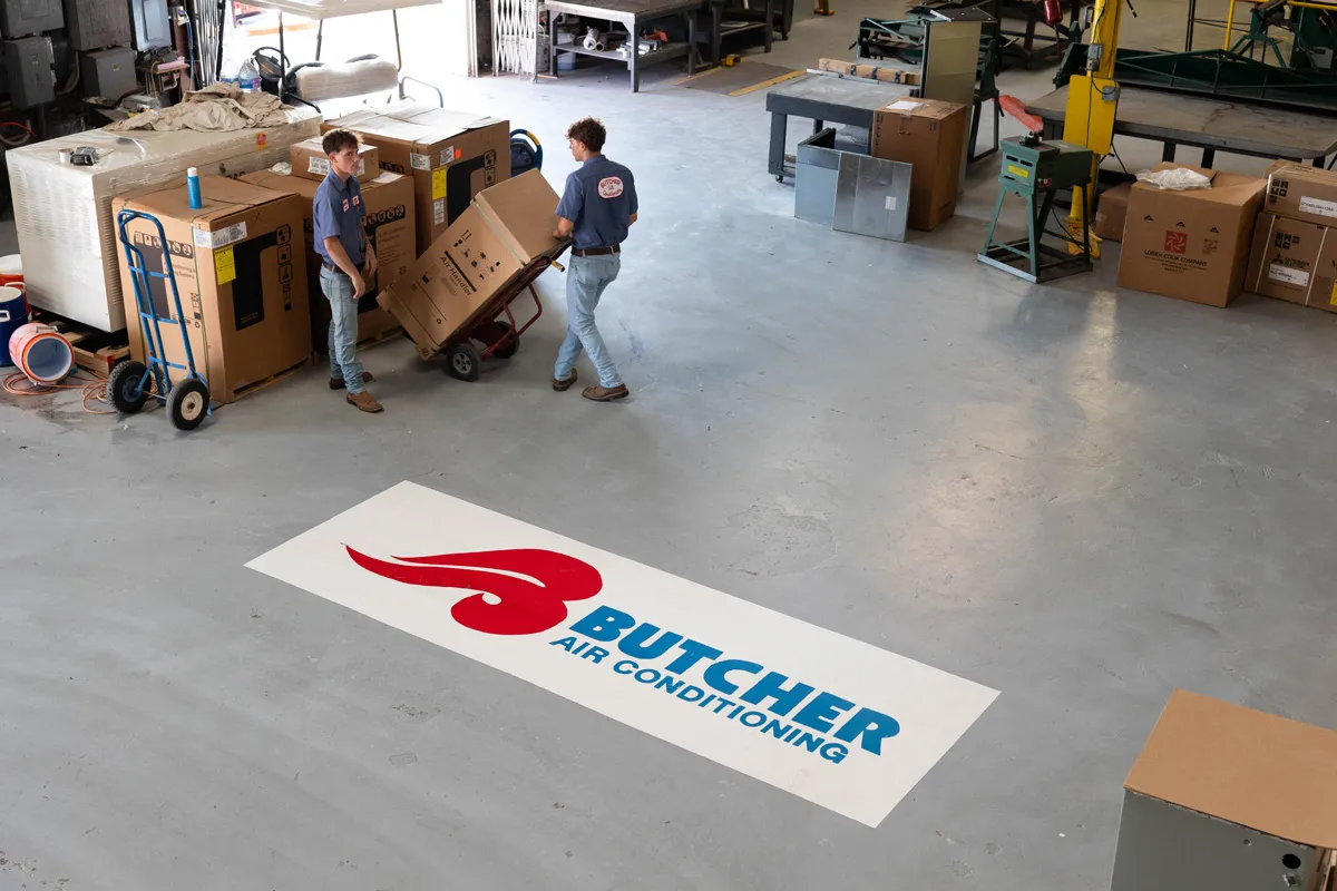 Two technicians moving packaged HVAC units in warehouse beside large Butcher Air Conditioning floor logo