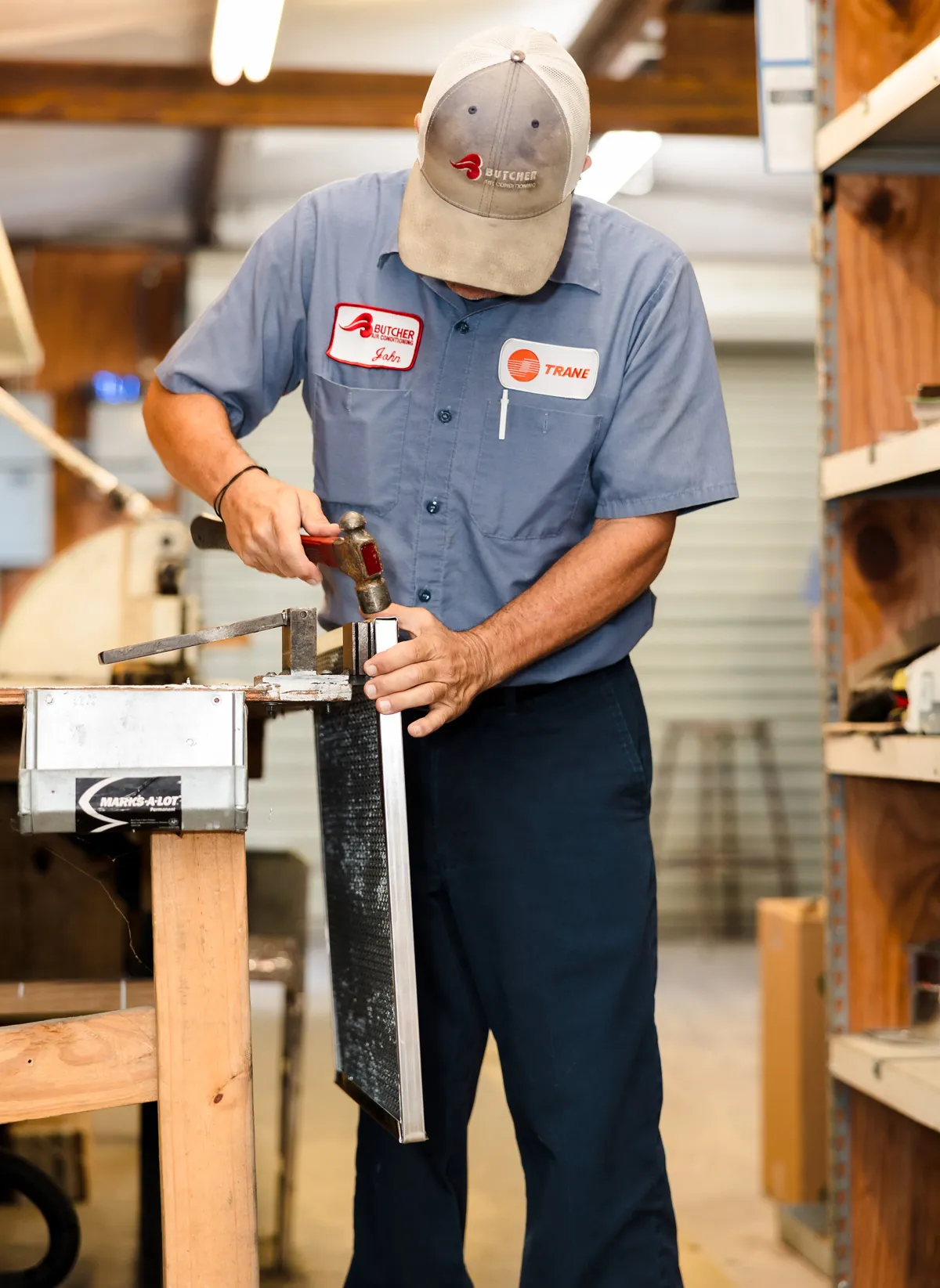 A man HVAC technician repairing an air conditioner filter with a hammer in a workshop, wearing a Trane uniform.