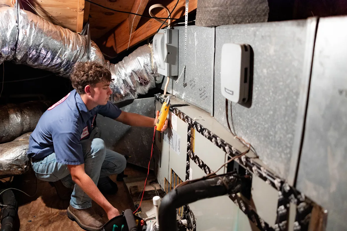 A man in an attic testing HVAC unit with a multimeter during furnace maintenance inspection
