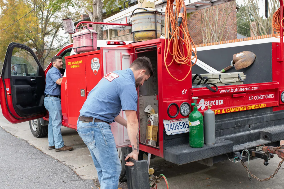 Two HVAC technicians loading equipment into red service truck beside sidewalk, gas cylinders and tools visible