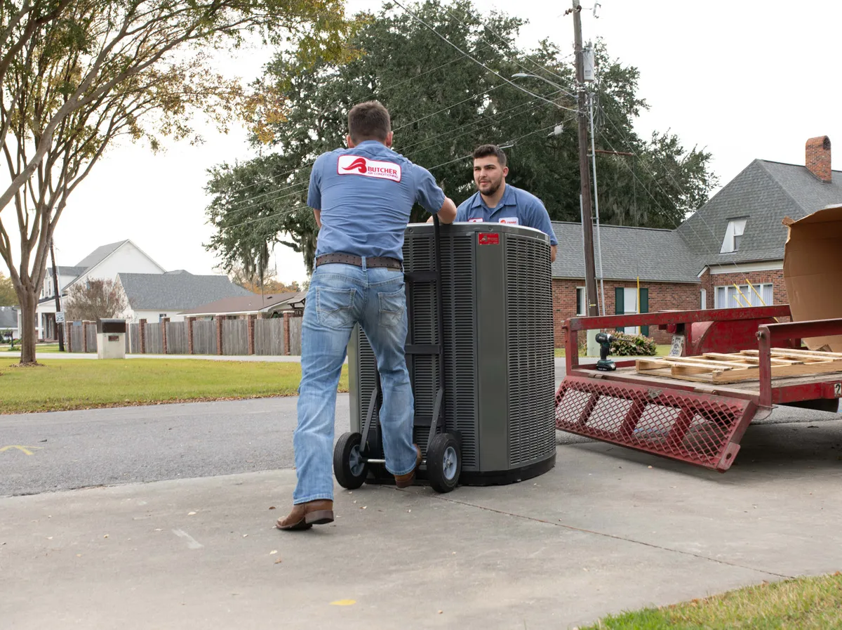 Two HVAC technicians moving an outdoor air conditioner unit on a dolly in a suburban driveway