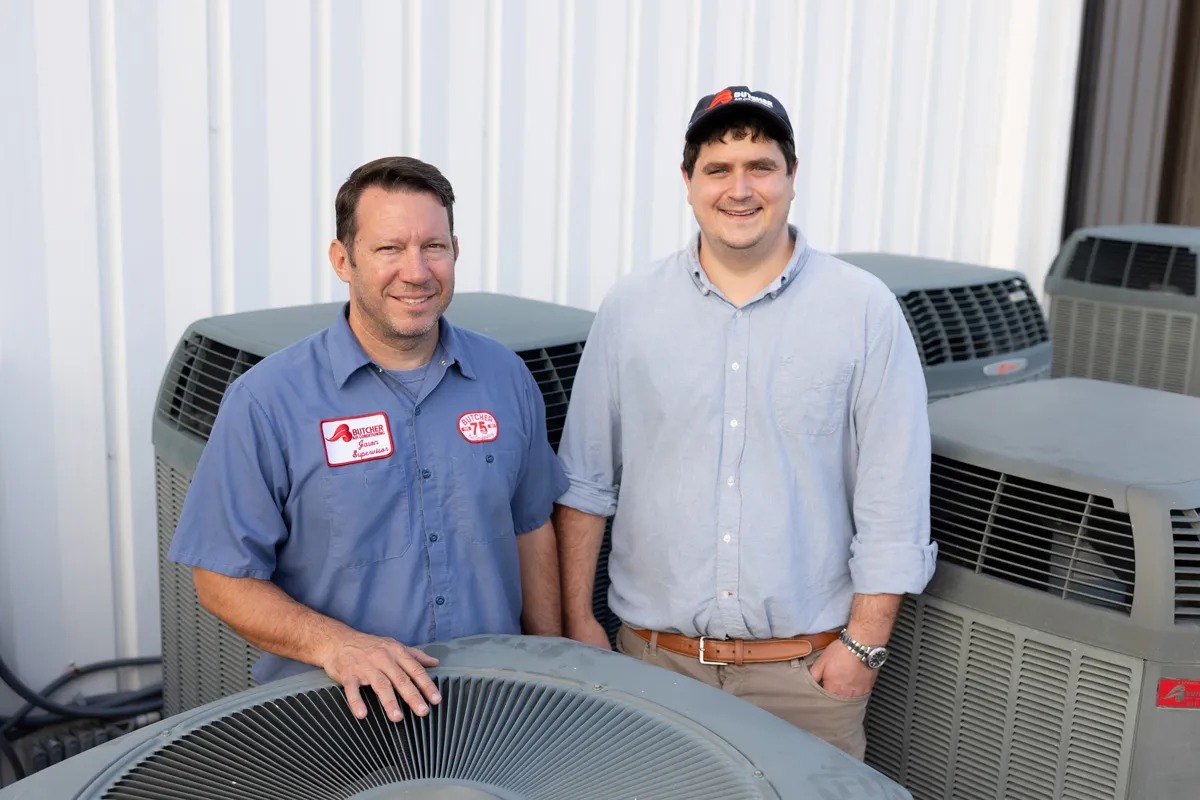 Two HVAC technicians standing beside commercial air conditioning units outside a metal building, smiling and posing